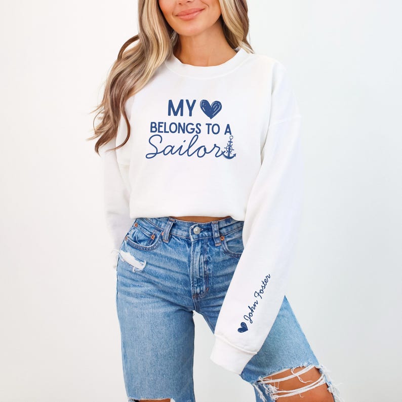 May include: White sweatshirt with the text "MY BELONGS TO A Sailor" in navy blue script. The sleeve has the name "John Foster" in navy blue script. The sweatshirt is paired with blue jeans.