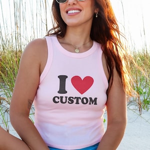 May include: A light pink tank top with the text "I &hearts; CUSTOM" in black and red. The top is a sleeveless style, perfect for warm weather. The image is taken outdoors, with a beach and tall grass in the background.