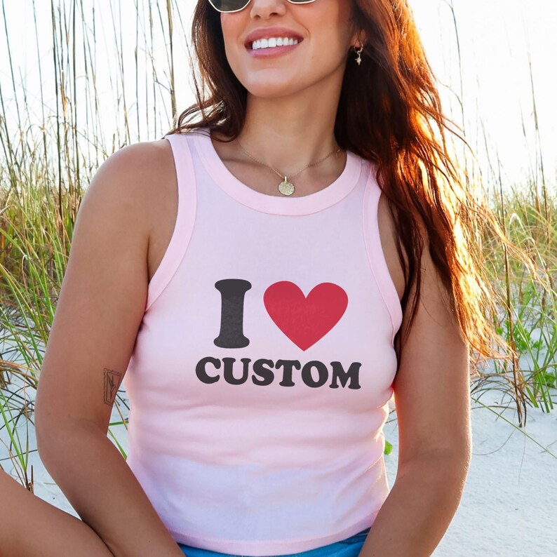 May include: A light pink tank top with the text "I &hearts; CUSTOM" in black and red. The top is a sleeveless style, perfect for warm weather. The image is taken outdoors, with a beach and tall grass in the background.
