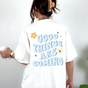 Puede incluir: Camiseta blanca con la frase "GOOD THINGS ARE COMING" en azul claro, con detalles de flores y estrellas. La camiseta es de estilo casual, adecuada para el uso diario.