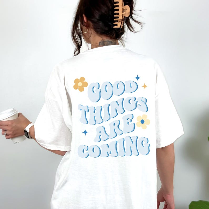 Puede incluir: Camiseta blanca con la frase "GOOD THINGS ARE COMING" en azul claro, con detalles de flores y estrellas. La camiseta es de estilo casual, adecuada para el uso diario.