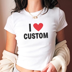 May include: White crew neck t-shirt with the text "I &hearts; CUSTOM" in black and a red heart. The shirt is short-sleeved and appears to be made of a soft material. A gold necklace is visible.