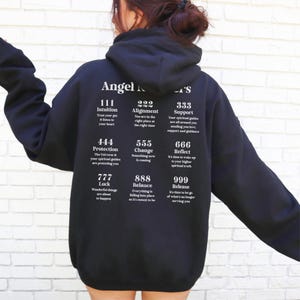 May include: Black hooded sweatshirt with white text. The text includes the words "Angel Numbers" and various number combinations with corresponding affirmations. The sweatshirt is a casual, comfortable style.