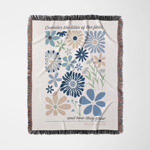 May include: Woven throw blanket with a floral design in shades of blue, beige, and gray. The blanket features the text "Consider the lilies of the field, and how they grow" and "Matthew 6:28". The edges are fringed.