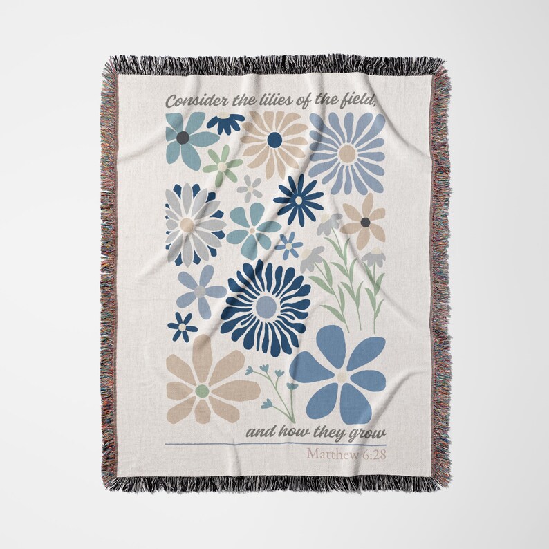 May include: Woven throw blanket with a floral design in shades of blue, beige, and gray. The blanket features the text "Consider the lilies of the field, and how they grow" and "Matthew 6:28". The edges are fringed.