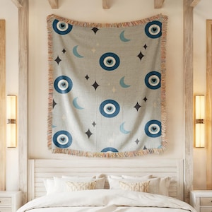 May include: A light gray woven throw blanket with a repeating pattern of blue eye symbols, crescent moons, and star shapes. The blanket has a fringed edge with orange, red, and tan threads. The blanket is hanging above a white bed.