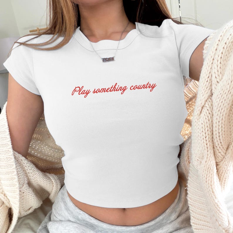 May include: A white short-sleeved ribbed t-shirt featuring the text "Play something country" printed in a red cursive font across the chest. The shirt has a crew neckline and a fitted silhouette, paired with a beige cable-knit cardigan.
