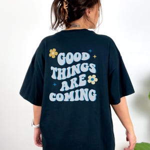 Puede incluir: Camiseta azul marino con la frase "GOOD THINGS ARE COMING" en blanco, azul claro y amarillo. El texto est&aacute; decorado con gr&aacute;ficos de flores y estrellas. La camiseta es de estilo casual y oversize.
