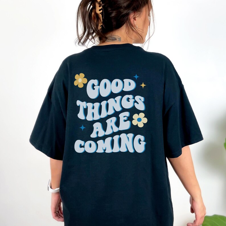 Puede incluir: Camiseta azul marino con la frase "GOOD THINGS ARE COMING" en blanco, azul claro y amarillo. El texto est&aacute; decorado con gr&aacute;ficos de flores y estrellas. La camiseta es de estilo casual y oversize.