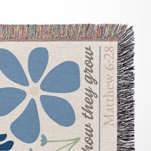 May include: Woven throw blanket with a large blue flower design and the text "how they grow Matthew 6:28". The blanket has a cream background with blue, green, and beige accents. The edges are fringed with multiple colors.