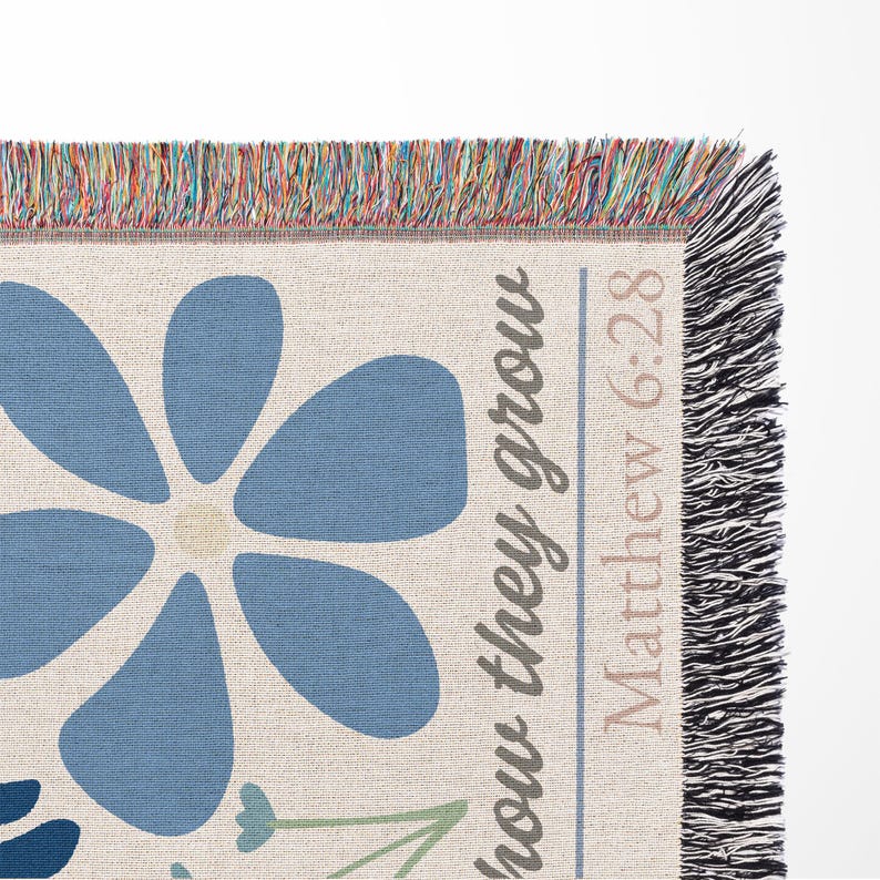 May include: Woven throw blanket with a large blue flower design and the text "how they grow Matthew 6:28". The blanket has a cream background with blue, green, and beige accents. The edges are fringed with multiple colors.