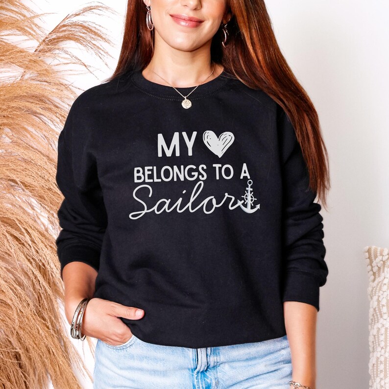 May include: Black crew neck sweatshirt with the text "MY HEART BELONGS TO A Sailor" in white. The design includes a heart and an anchor. The sweatshirt is a solid black color.