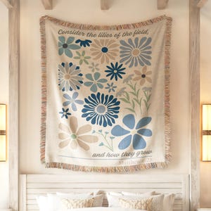 May include: A cream-colored throw blanket with a floral design in shades of blue, gray, and beige. The blanket features the text "Consider the lilies of the field, and how they grow, Matthew 6:28."