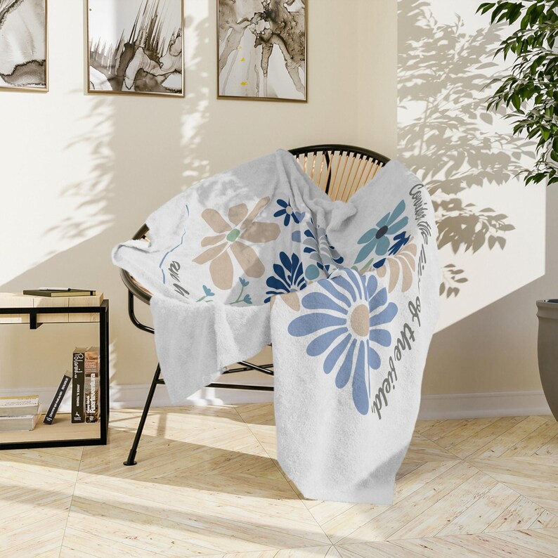 May include: White throw blanket with large blue, tan, and navy floral designs. The blanket is draped over a black wicker chair. Text on the blanket reads "Consider the lilies of the field."
