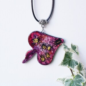 May include: A unique pendant necklace featuring a purple, leaf-shaped pendant with gold-colored beetle designs. The pendant hangs from a black cord with a silver-colored metal clasp. The background is white, with green and white ivy leaves in the lower right corner.
