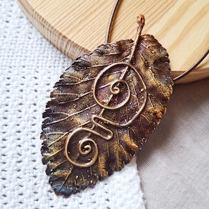 May include: A handcrafted leaf pendant with a bronze finish and intricate wire detailing. The pendant features a spiral design and is suspended from a thin, brown cord. The leaf has a textured surface with shades of brown, gold, and hints of blue.