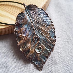 May include: A handcrafted leaf pendant with a detailed, textured surface. The pendant features a copper-colored stem and a decorative swirl. The leaf is primarily bronze and blue, with hints of purple and green. It hangs from a black cord.