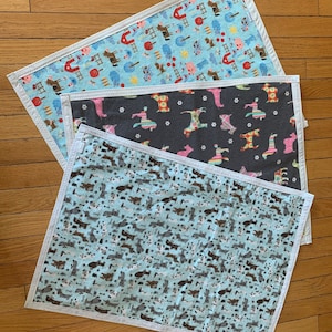May include: Three different fabric squares with white borders. The top square is light blue with a white background and a pattern of horses, barns, and red apples. The middle square is dark gray with a white background and a pattern of dogs. The bottom square is light blue with a white background and a pattern of dogs.