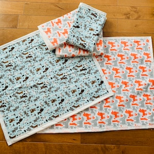 May include: Four different fabric squares with a pattern of dogs in various colors. The squares are arranged in a stack, with the largest square on the bottom and the smallest square on top. The squares are all made of a soft, cotton fabric.