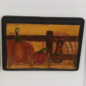 May include: A rectangular, framed painting with a black border. The artwork depicts a fall scene with pumpkins, a turkey, and a fence. The pumpkins are orange and red, and the turkey is brown. The background is yellow.