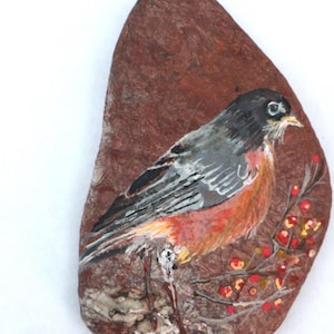 May include: Hand-painted rock art featuring a robin perched on a branch with red berries. The robin is painted in shades of gray, black, orange, and white. The background is a brown rock with a rough texture.