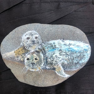 May include: A hand-painted rock depicting two grey seals, one on top of the other, with a blue and white background resembling water.