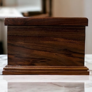 Op de afbeelding: Een donkerbruine houten doos met een eenvoudig ontwerp. De doos heeft een gladde, gepolijste afwerking en een licht verhoogde basis.