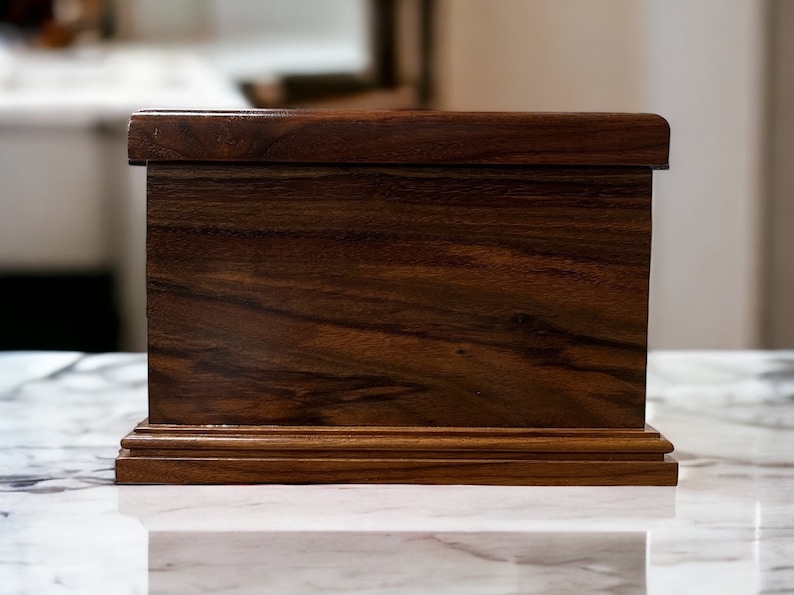 Op de afbeelding: Een donkerbruine houten doos met een eenvoudig ontwerp. De doos heeft een gladde, gepolijste afwerking en een licht verhoogde basis.