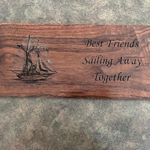 Op de afbeelding: Een houten plaquette met een gegraveerd beeld van een zeilboot en de tekst "Best Friends Sailing Away Together".