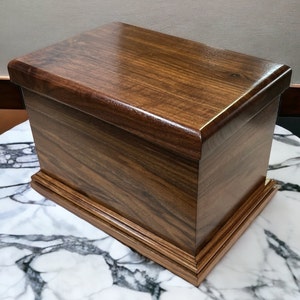Op de afbeelding: Een donkerbruine houten doos met een scharnierend deksel en een decoratieve rand rond de basis. De doos is gemaakt van een rijk hout met een natuurlijk houtnerfpatroon.