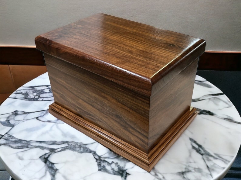 Op de afbeelding: Een donkerbruine houten doos met een scharnierend deksel en een decoratieve rand rond de basis. De doos is gemaakt van een rijk hout met een natuurlijk houtnerfpatroon.