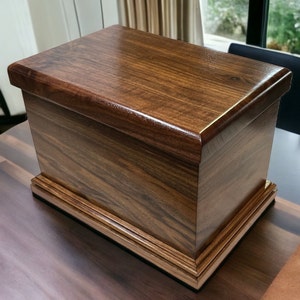 Op de afbeelding: Een donkerbruine houten doos met een scharnierend deksel en een decoratieve rand rond de basis. De doos is gemaakt van massief hout en heeft een gladde, gepolijste afwerking.