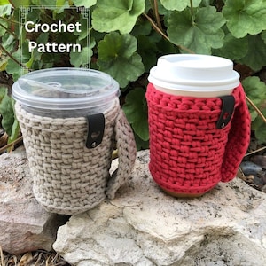 Peut inclure: Deux manchons à café en crochet, l'un beige et l'autre rouge, avec des lanières en cuir noir. Les manchons sont posés sur un rocher.