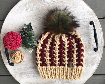 May include: A knitted hat with a burgundy and cream color pattern and a brown faux fur pom pom. The hat has a small brown leather tag with the text "RISB" on it.
