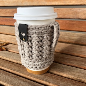 May include: A beige crocheted coffee cup cozy with a black leather handle and a gold heart-shaped button. The cozy is on a wooden surface.