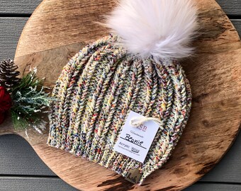 Może przedstawiać: Kolorowa dzianinowa czapka z białym pomponem. Czapka jest wykonana z akrylowej wełny i ma metkę z napisem "Handmade Beanie Acrylic Wool".