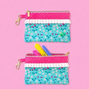 May include: Two small, rectangular pouches with gold zippers and key ring clasps. The top section is pink with white polka dots, and the bottom is turquoise with a floral pattern. A white ruffle separates the two sections. One pouch is open, revealing colorful markers.