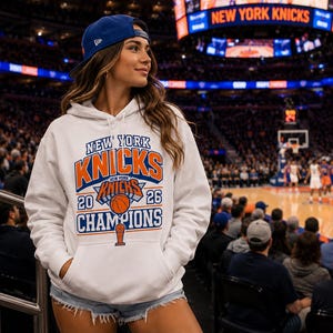May include: A white hooded sweatshirt featuring a graphic print that reads New York Knicks 2026 Champions with an illustration of a basketball and a trophy. The hoodie has a front kangaroo pocket and white drawstrings at the neckline.