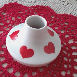 May include: A small, white ceramic vase with red heart designs. The vase sits on a red, crocheted doily, with a white lace tablecloth in the background. The vase has a cylindrical neck and a rounded base.