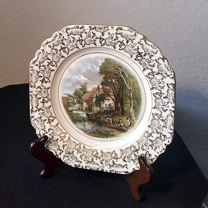 May include: A white ceramic plate with a gold rim and a black and white floral design. The center of the plate features a colorful image of a cottage with a river and trees.