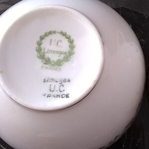 May include: A white porcelain saucer with a rounded edge. The underside features a green wreath design with the text "U.C. Limoges FRANCE". The saucer is part of a tableware set.