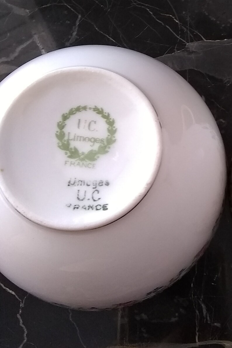 May include: A white porcelain saucer with a rounded edge. The underside features a green wreath design with the text "U.C. Limoges FRANCE". The saucer is part of a tableware set.