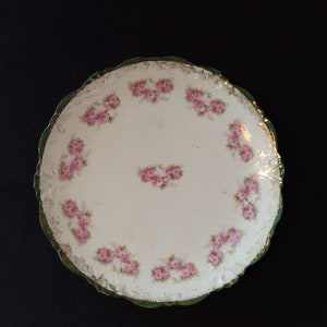 May include: A white porcelain plate with a green and gold rim. The plate is decorated with a delicate pattern of pink roses.
