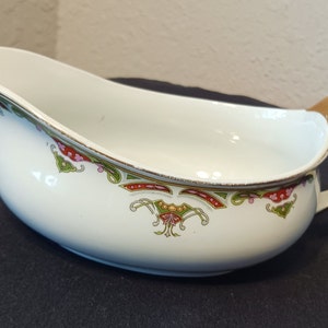 May include: A white porcelain gravy boat with a gold rim and a floral design in shades of green, red, and pink. The handle is also gold.