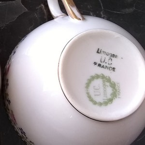 May include: White porcelain teacup with a gold handle and floral detailing. The bottom of the cup features the text "Limoges - U.C. FRANCE" within a green wreath. A classic piece for tea or coffee.