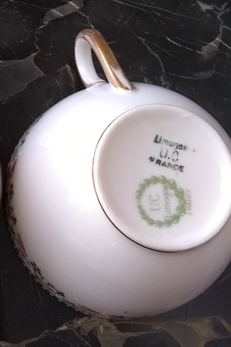 May include: White porcelain teacup with a gold handle and floral detailing. The bottom of the cup features the text "Limoges - U.C. FRANCE" within a green wreath. A classic piece for tea or coffee.