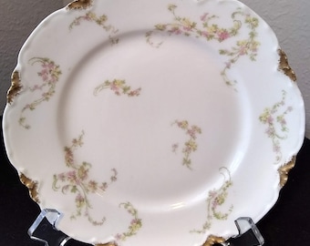 May include: A white porcelain dinner plate with a scalloped edge and a gold trim. The plate features a delicate floral pattern in shades of pink and green.
