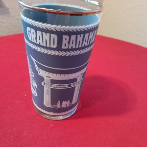 May include: A blue glass tumbler with a white rim and a white design that says "Grand Bahama International Bazaar".