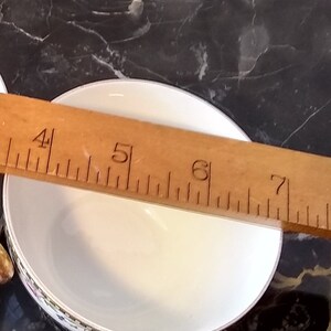 May include: A white ceramic bowl with a decorative floral border, approximately 6 inches in diameter, is measured with a wooden ruler. The bowl sits on a dark, marbled surface.