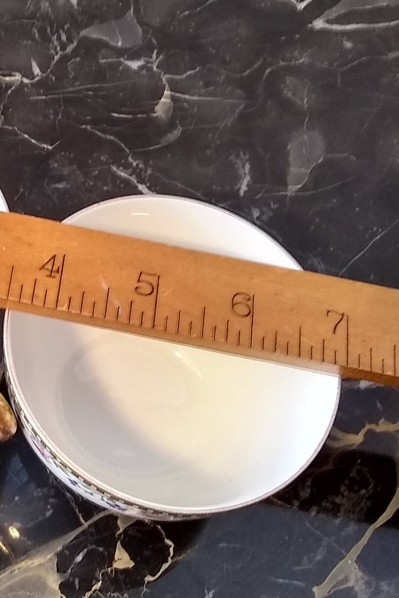 May include: A white ceramic bowl with a decorative floral border, approximately 6 inches in diameter, is measured with a wooden ruler. The bowl sits on a dark, marbled surface.